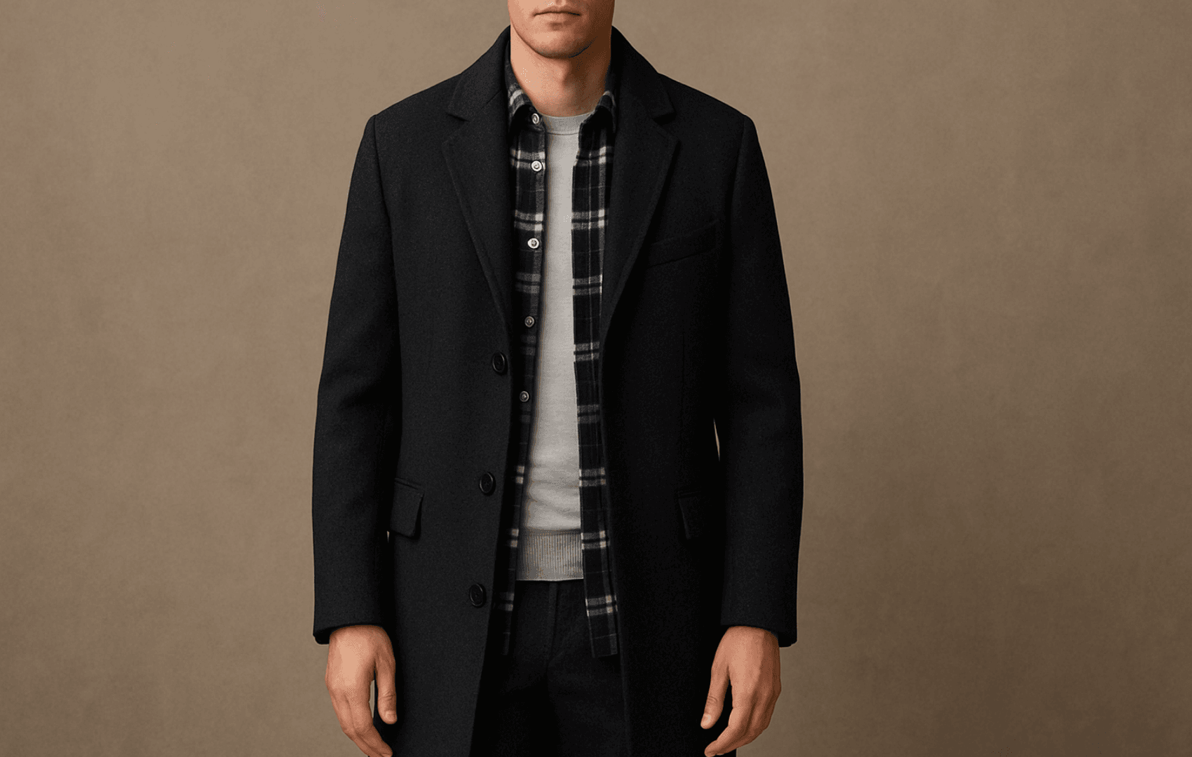 A man facing forward in a charcoal wool topcoat layered over a navy-and-white plaid flannel shirt and light gray merino tee, shown from chest to thigh against a neutral background.