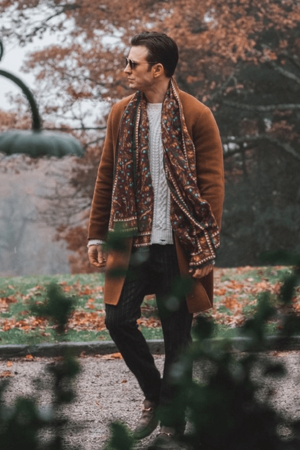 Chris Evans standing in an elegant vintage study wearing a rust-orange wool overcoat layered over a light beige crew-neck sweater. A multicolor patterned scarf featuring red, green, and blue tones adds contrast, while navy pinstripe trousers and brown leather loafers complete the look. The warm, book-filled background enhances the classic autumn fashion aesthetic.