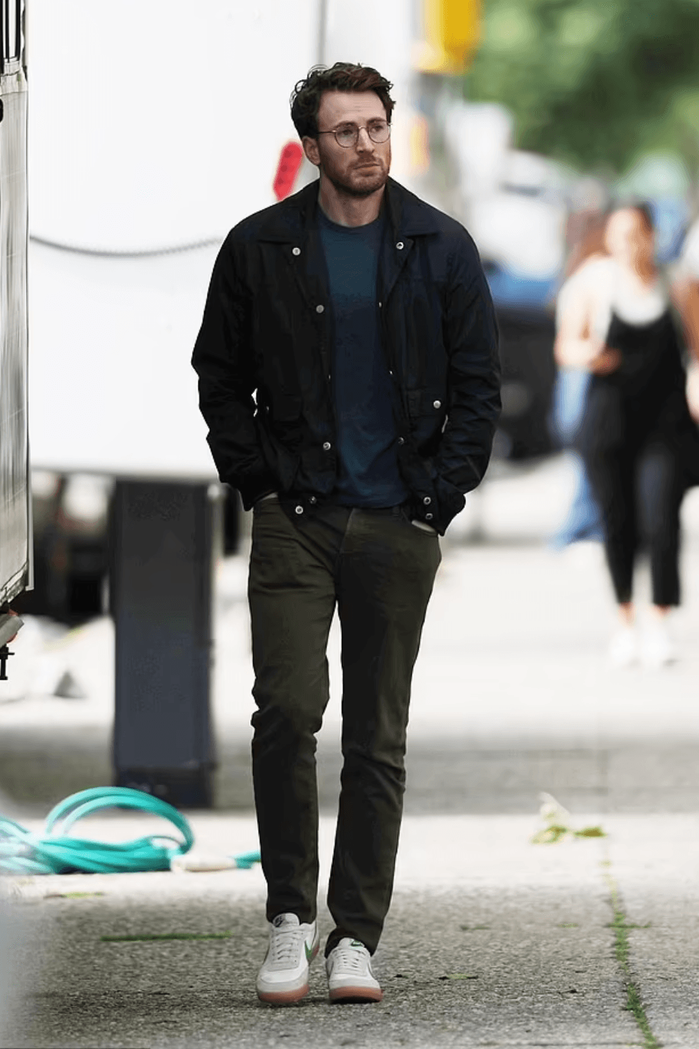 Chris Evans walking outdoors in casual streetwear — a tan suede bomber jacket over a plain white T-shirt, paired with dark green chinoss, white sneakers, dark sunglasses. He’s captured mid-stride on a city sidewalk, exuding relaxed, confident style.
