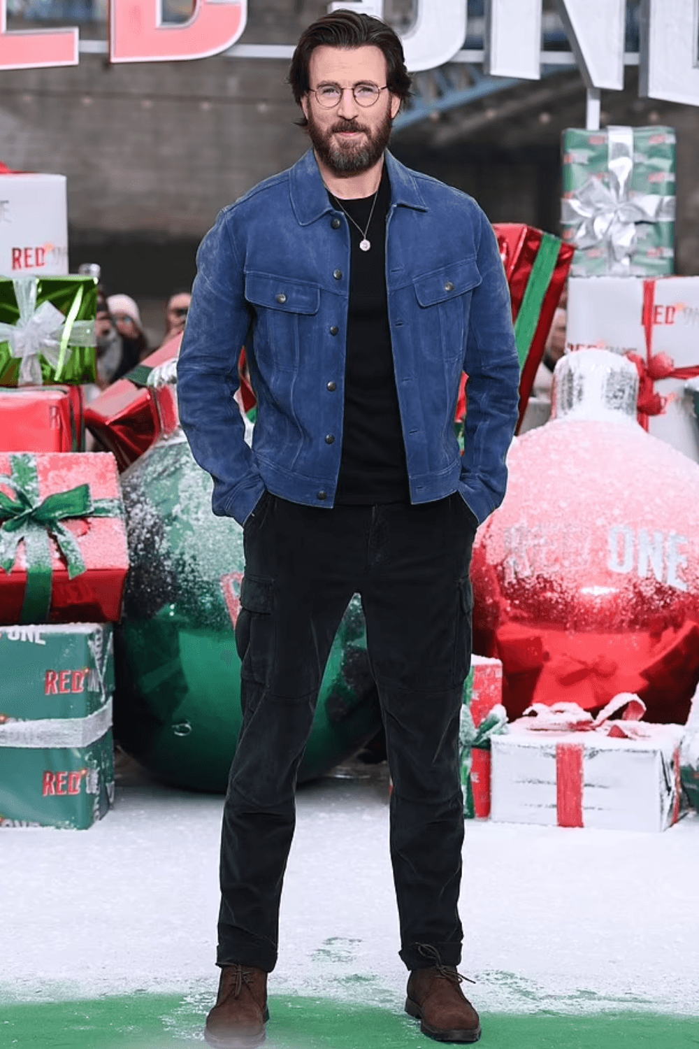 Chris Evans wearing a blue suede trucker jacket over a black T-shirt, styled with black cargo trousers, brown suede boots, and a simple pendant necklace, posing outdoors on a festive film premiere backdrop.