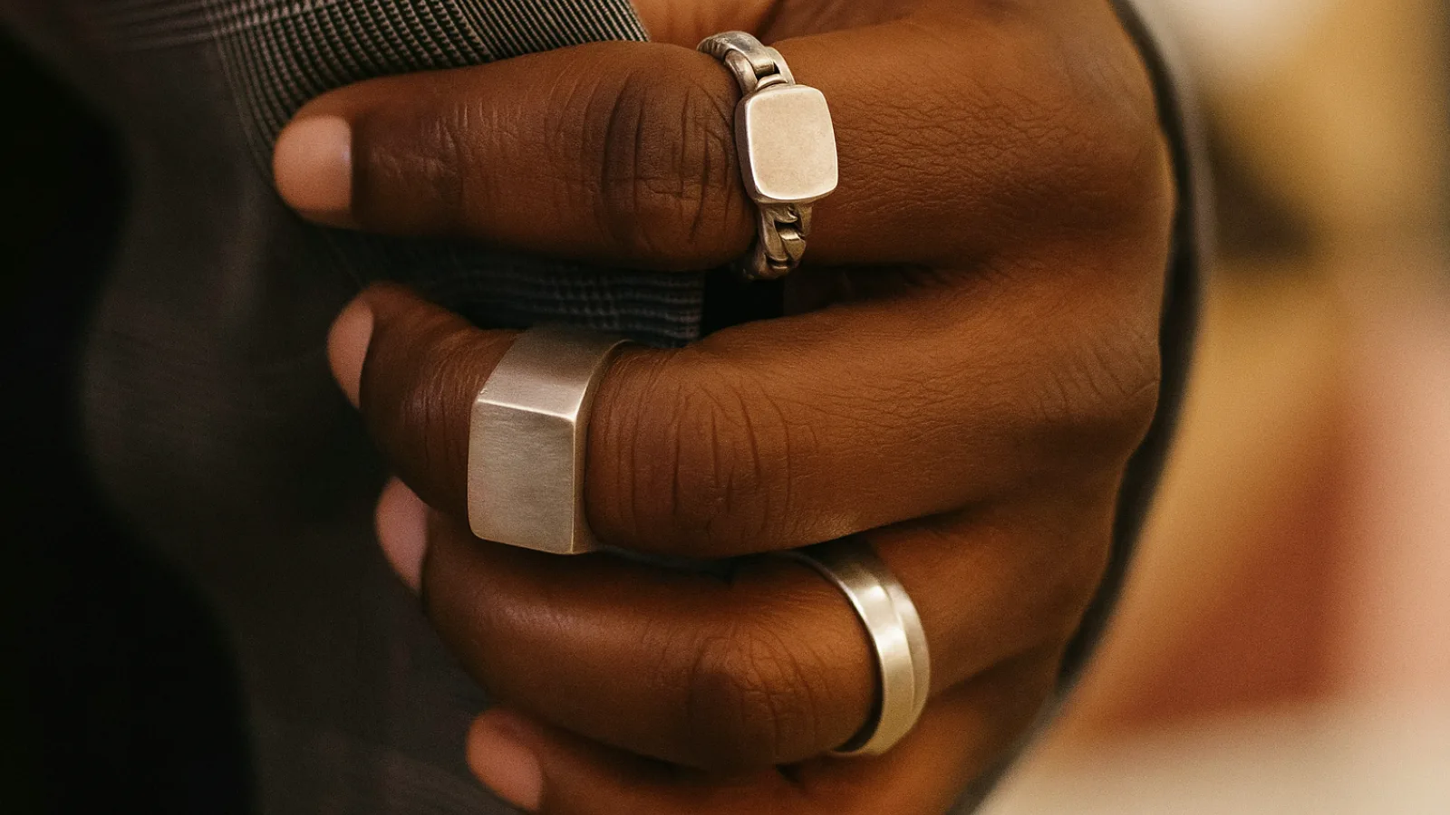 Close-up of a man’s hand with warm brown skin gripping a checkered suit lapel, wearing three silver rings: a chain-link ring on the index finger, a square-faced signet ring on the middle finger, and a polished silver band on the ring finger. The background is softly blurred with warm lighting, highlighting the metal’s realistic texture.