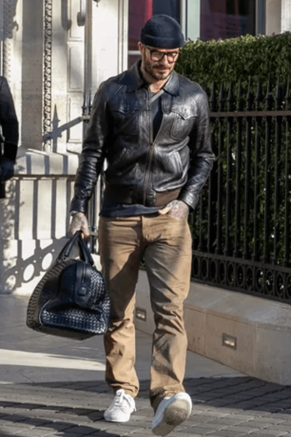 David Beckham walks down a city street wearing a black leather jacket, gray beanie, sunglasses, khaki pants, and white sneakers while carrying a large black leather bag.