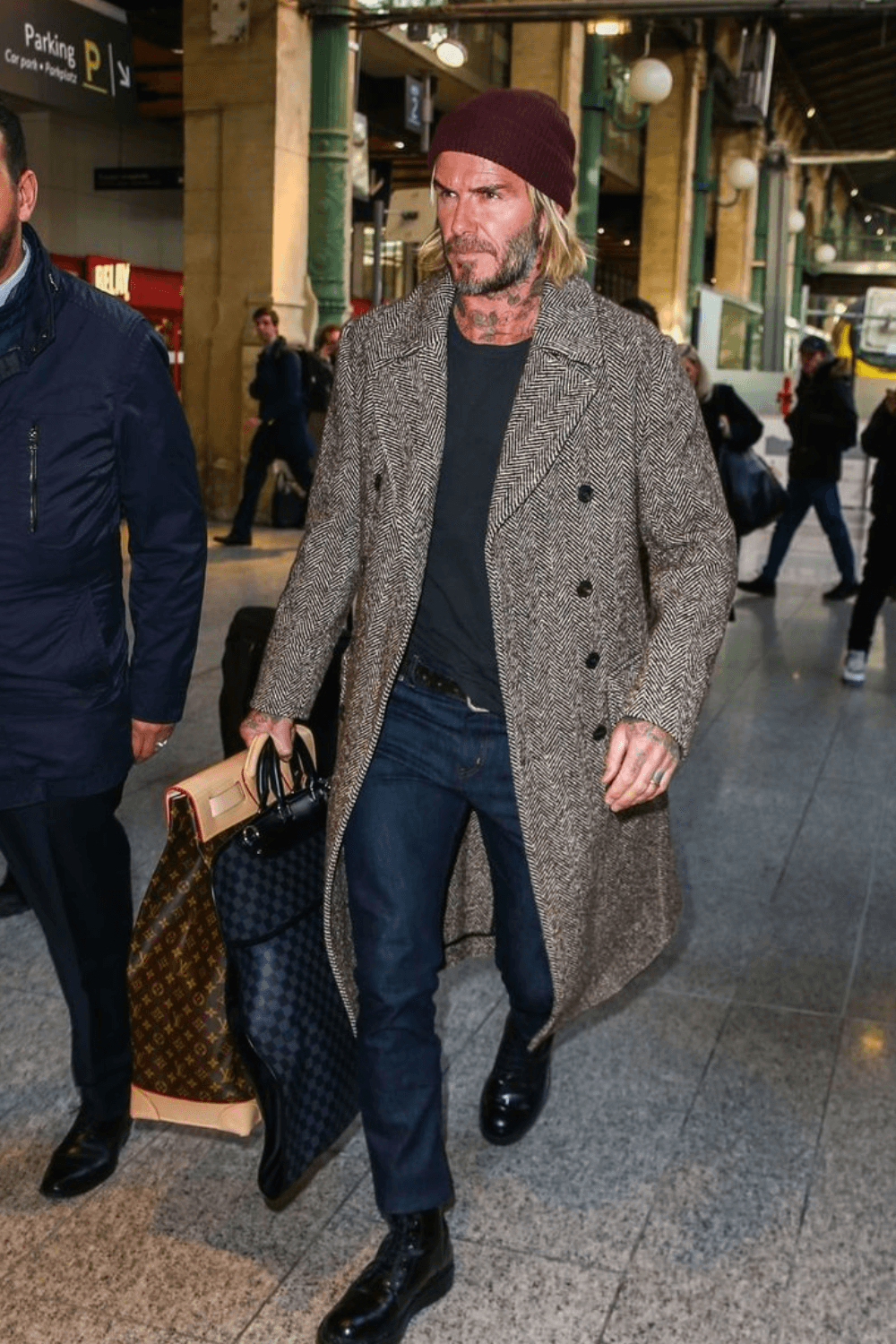 David Beckham wearing a long tweed overcoat layered over a dark sweater and jeans, paired with a burgundy knit beanie and dark boots, walking in an urban setting.