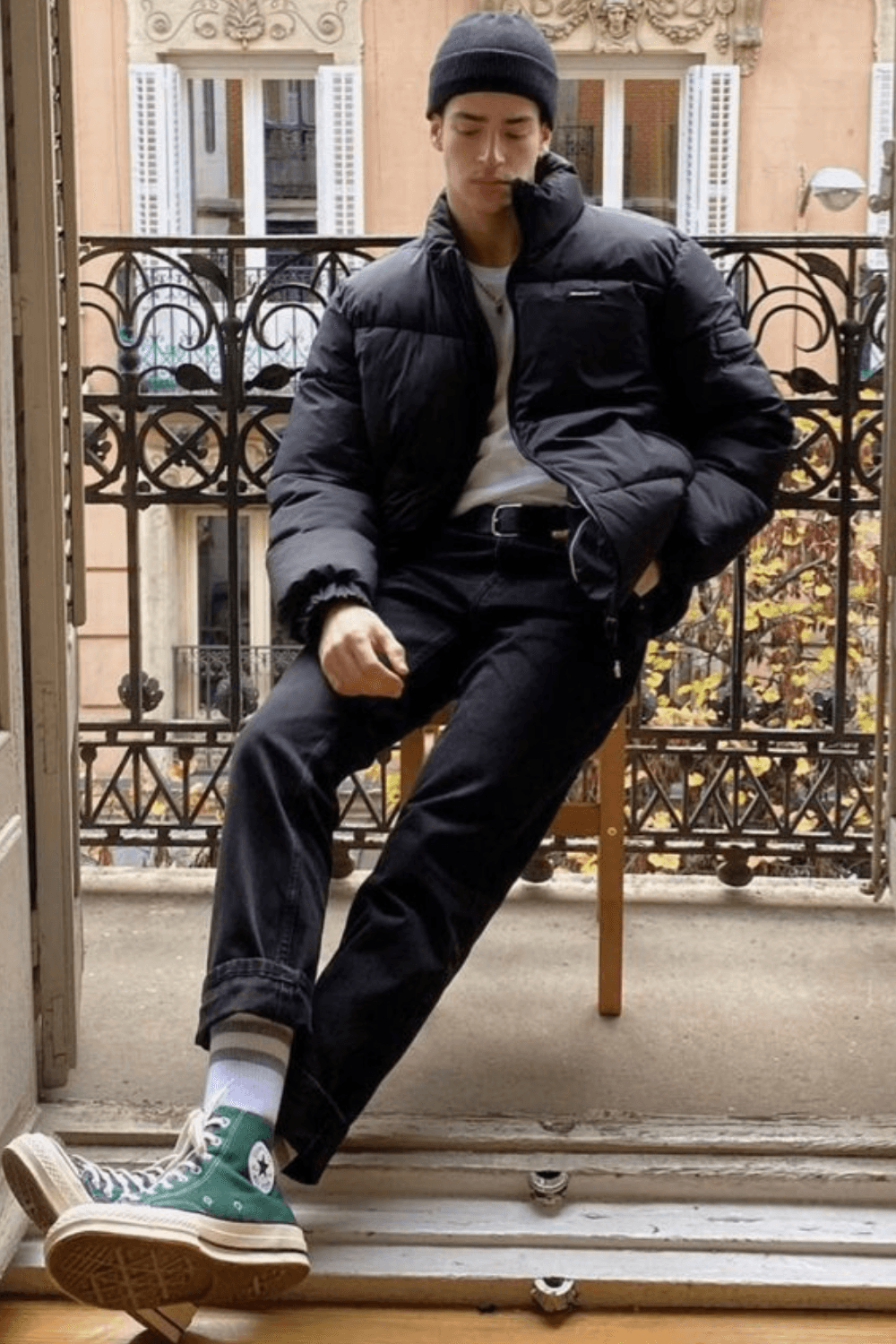 Manu Rios sitting on a balcony wearing a black puffer jacket over a white T-shirt, black jeans, white socks, and green high-top Converse sneakers, accessorized with a black beanie.