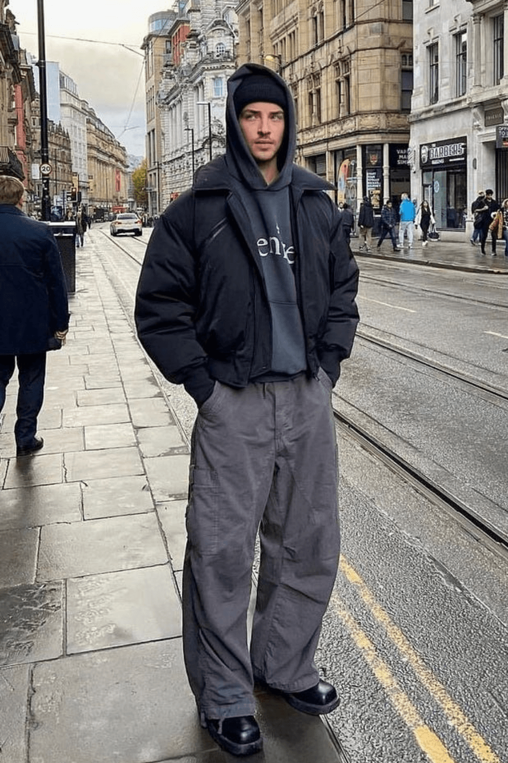 Manu Rios standing on a city street wearing an oversized black puffer jacket layered over a charcoal gray hoodie, wide-leg gray cargo pants, and black leather shoes, styled with a black beanie.