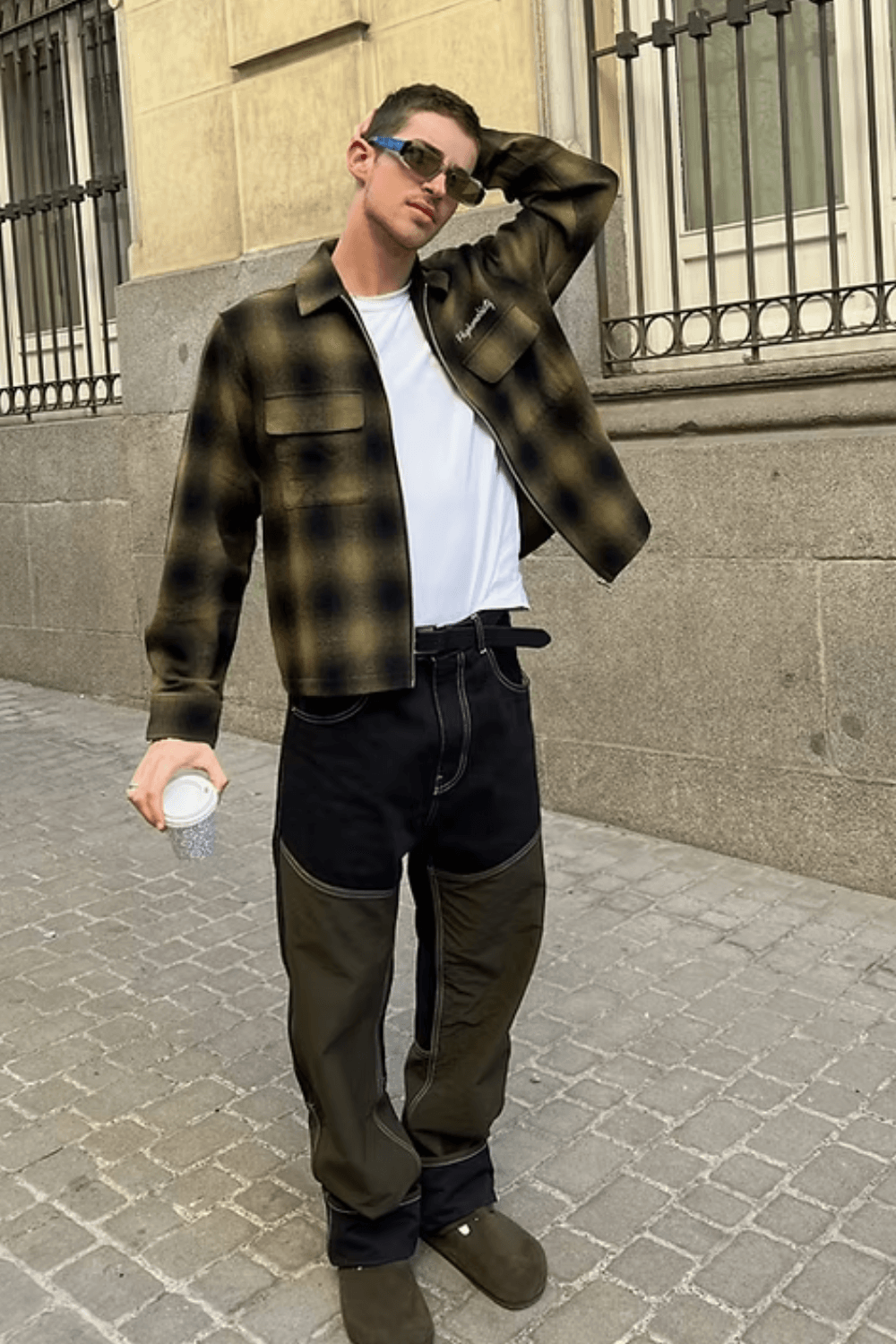 Manu Rios wearing a cropped yellow-and-black plaid overshirt layered over a plain white T-shirt, paired with two-tone black and olive cargo jeans and brown suede clogs, accessorized with sunglasses and a coffee cup.