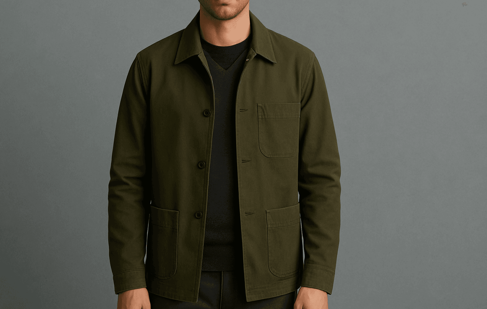 A man wearing a dark olive cotton chore coat layered over a charcoal V-neck wool sweater with a hidden black merino tee, shown from face to thighs against a neutral backdrop.