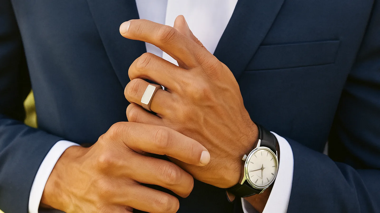 Close-up of a man’s hands adjusting a navy suit jacket, wearing a polished silver signet ring and a silver wristwatch with a black leather strap. The white shirt cuffs peek slightly under the sleeves, captured in natural daylight with a blurred outdoor background.