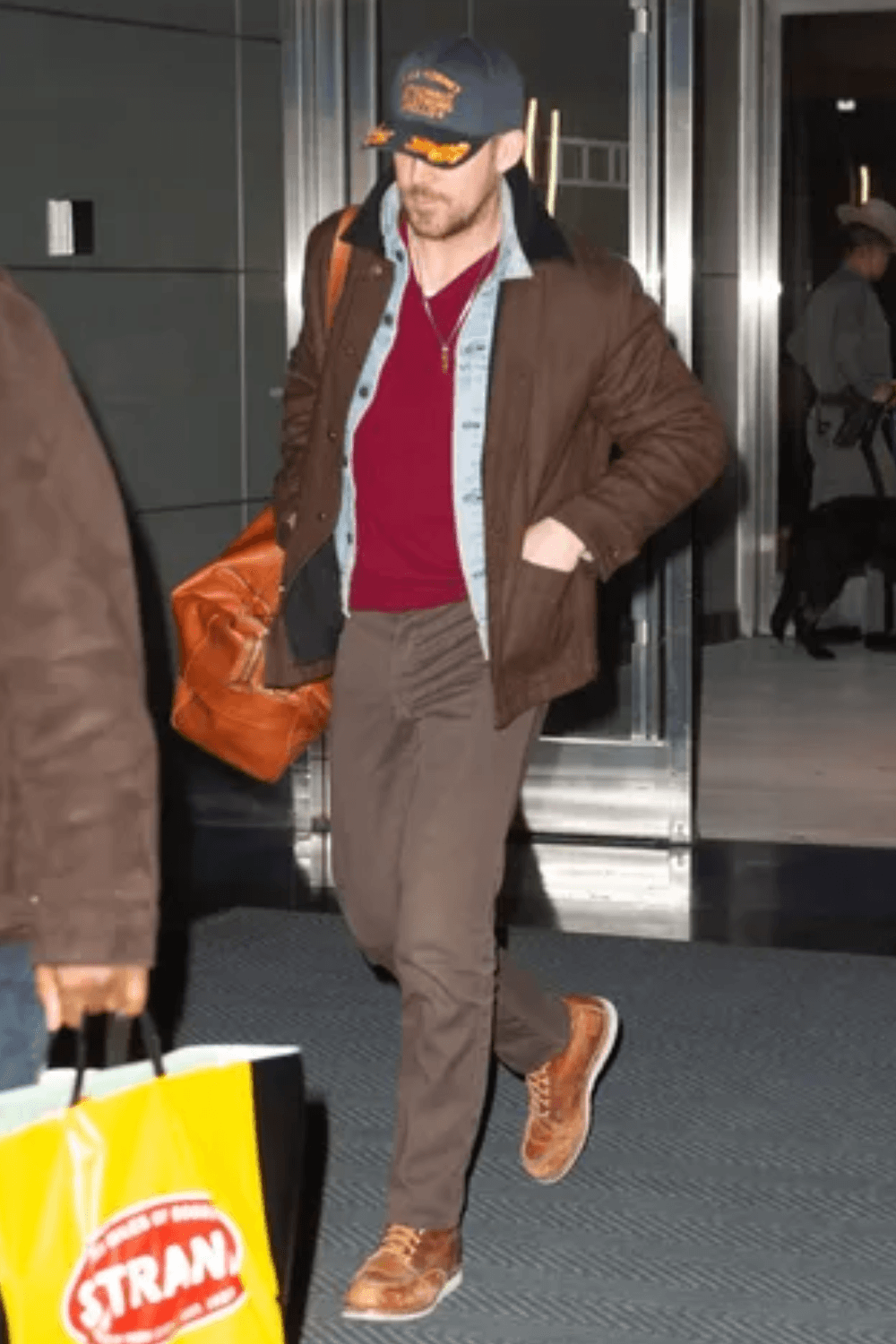 Ryan Gosling walking outdoors surrounded by people, dressed in a brown overshirt layered over a light denim jacket and red shirt, with brown pants and tan lace-up boots.