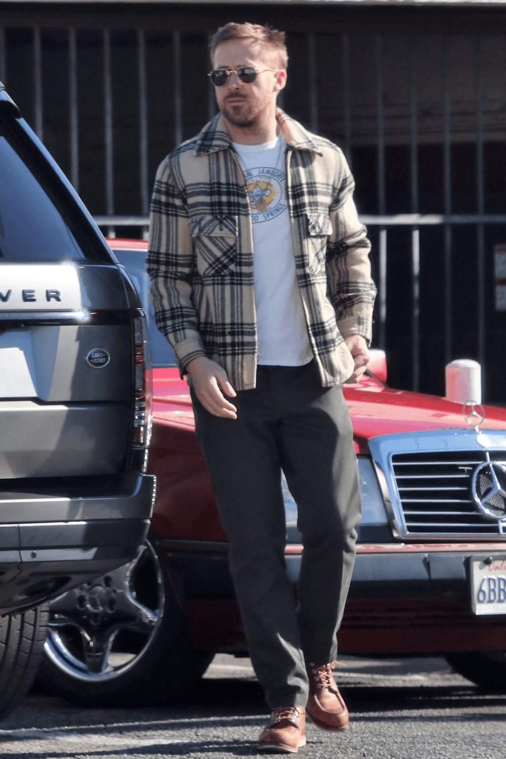 Ryan Gosling stepping outdoors on a sidewalk, dressed in a layered outfit — a brown checkered overshirt over a darker jacket, paired with black jeans and a light-colored baseball cap, walking past street scenes and storefronts.