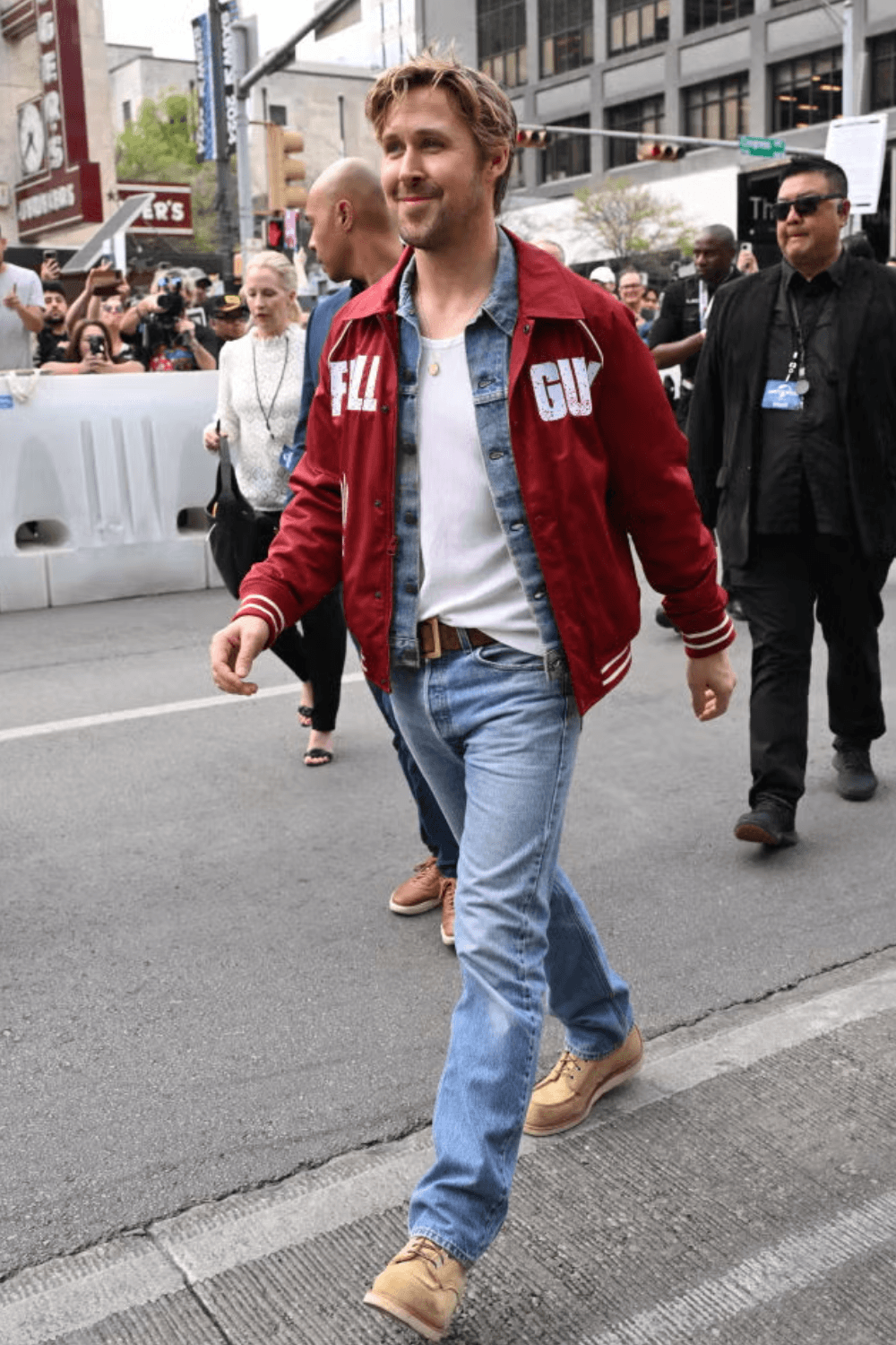 A medium shot of actor Ryan Gosling standing in front of a SXSW step-and-repeat backdrop, wearing a red jacket with “FALL GUY” printed, light blue shirt underneath, looking slightly off camera with a neutral expression.