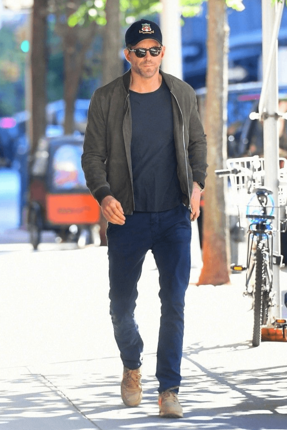 Ryan Reynolds walking outdoors on a city sidewalk, dressed in a dark suede bomber jacket, navy T-shirt, slim blue pants, tan lace-up boots, black sunglasses, and a black baseball cap, with sunlight and urban scenery in the background.