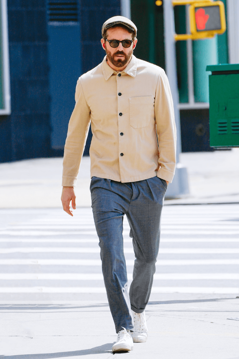 Ryan Reynolds walking across a city crosswalk wearing a beige button-up overshirt over a light top, gray pleated trousers, and white sneakers. Accessories include a tweed flat cap and dark round sunglasses. The outfit reflects smart-casual men’s street style with minimalist tones and subtle vintage influence.