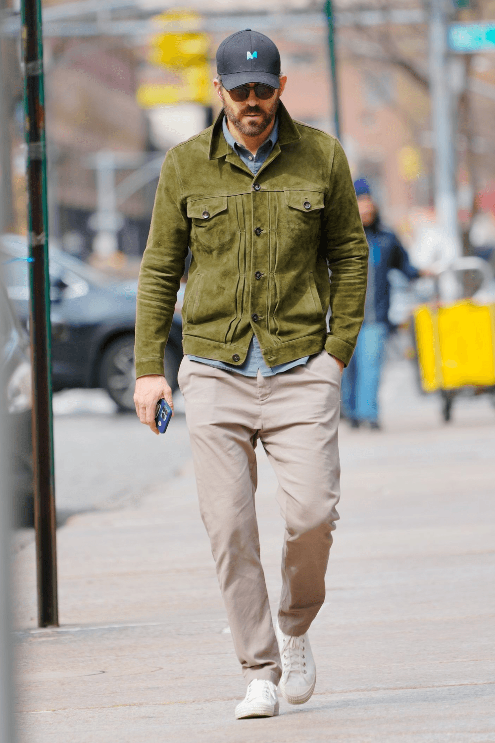 Ryan Reynolds walks through the city in a relaxed yet polished smart-casual outfit. He wears an olive green suede trucker jacket layered over a light chambray shirt, paired with beige chinos and crisp white sneakers. The look is finished with dark sunglasses and a black baseball cap, creating an effortlessly cool weekend ensemble that blends classic style with modern streetwear comfort.