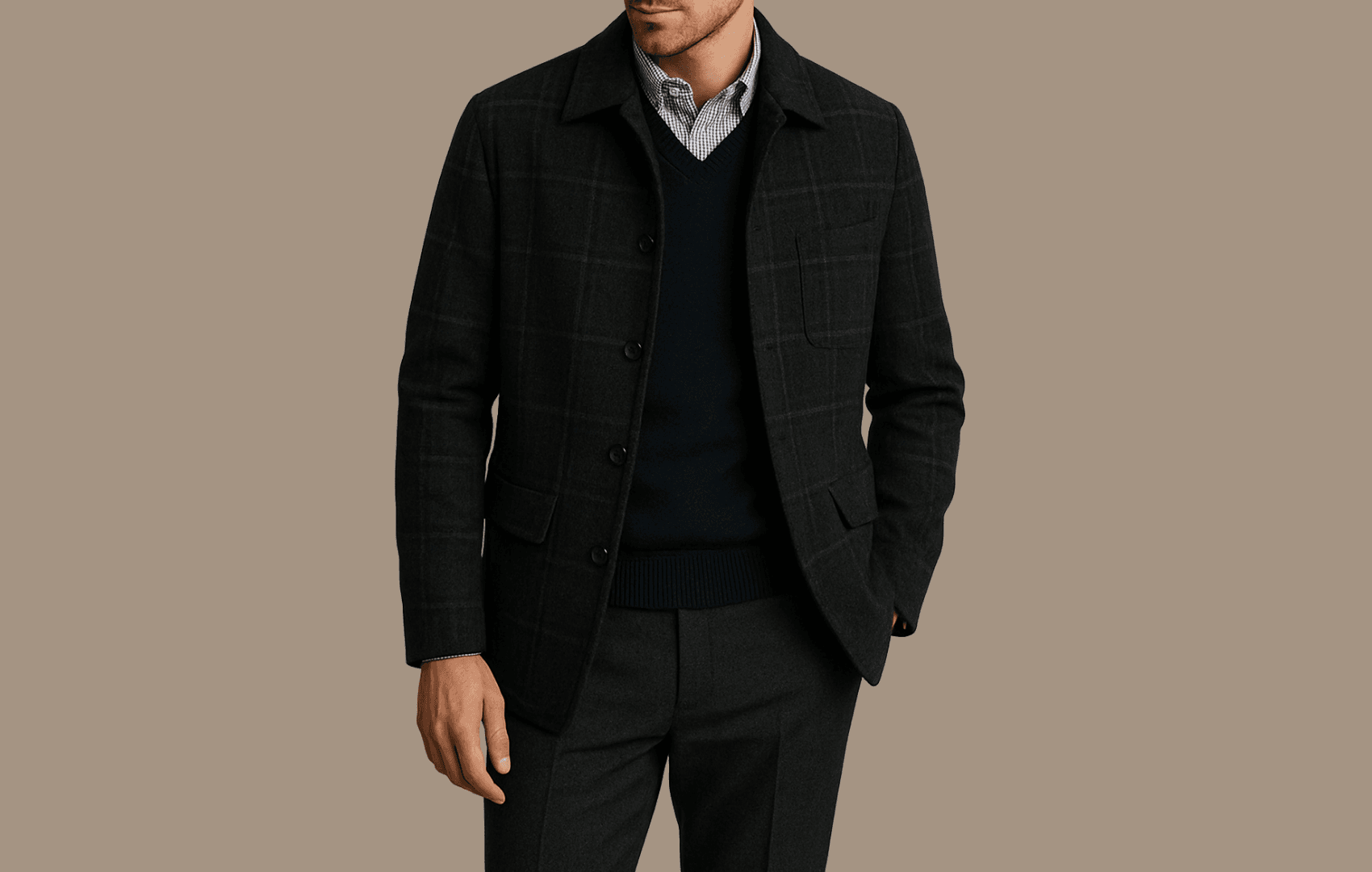 A stylish man wearing a gray striped Oxford shirt under a navy merino wool V-neck sweater and a dark charcoal plaid wool overshirt or sports coat, standing against a neutral background with soft lighting.
