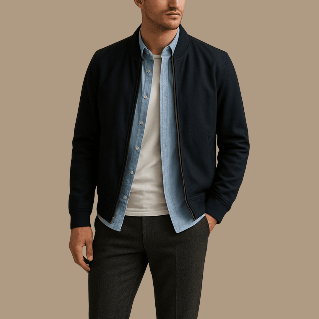 A stylish man wearing a navy bomber jacket layered over a light blue Oxford shirt and a white crew neck tee. He stands against a neutral beige background under soft lighting, highlighting the refined textures and relaxed layering.