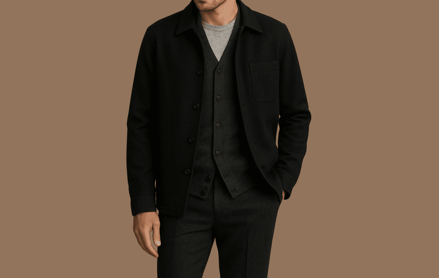 A stylish man wearing a monochromatic layered outfit: a light gray crew-neck tee, charcoal wool V-neck cardigan, and black overshirt, paired with dark gray trousers, standing against a neutral beige background under soft, editorial lighting.