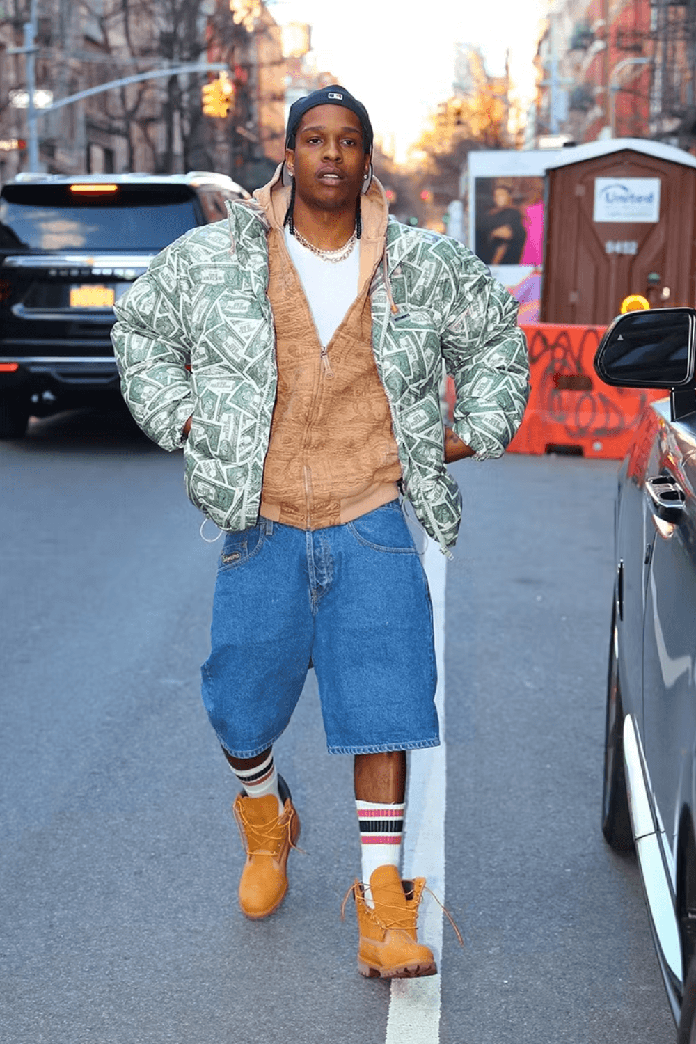 A$AP Rocky wearing a navy beanie, white T-shirt, brown jacket layered under a green puffer jacket, dark denim shorts, white socks, and brown shoes, standing with both hands in his jacket pockets.