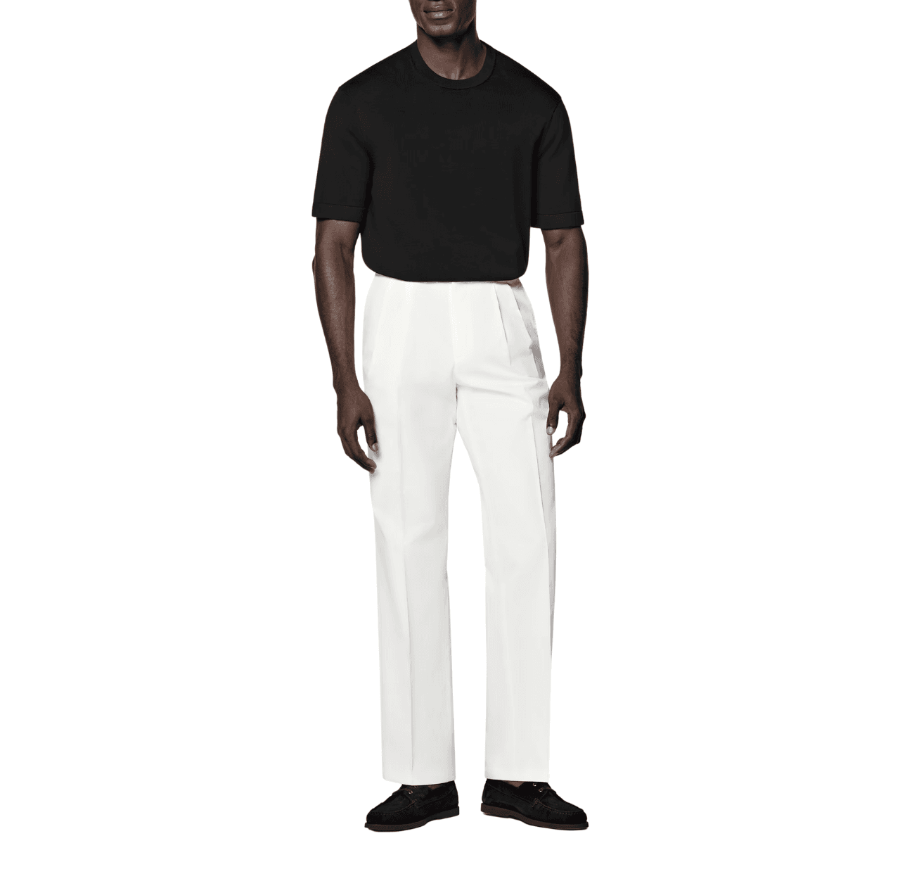 Man wearing a black T-shirt with white pants and black suede loafers, standing with both arms relaxed at his sides.