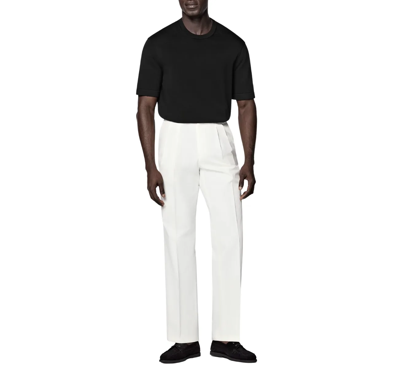 Man wearing a black T-shirt with white pants and black suede loafers, standing with both arms relaxed at his sides.