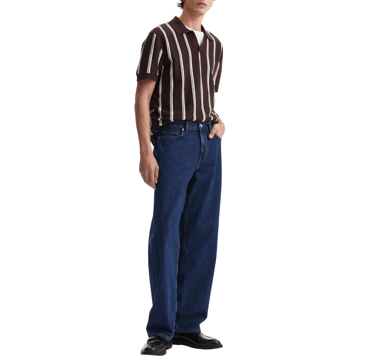 Man wearing a white tee, a brown striped knit polo, dark denim blue jeans, and black loafers, one hand in pants pocket.