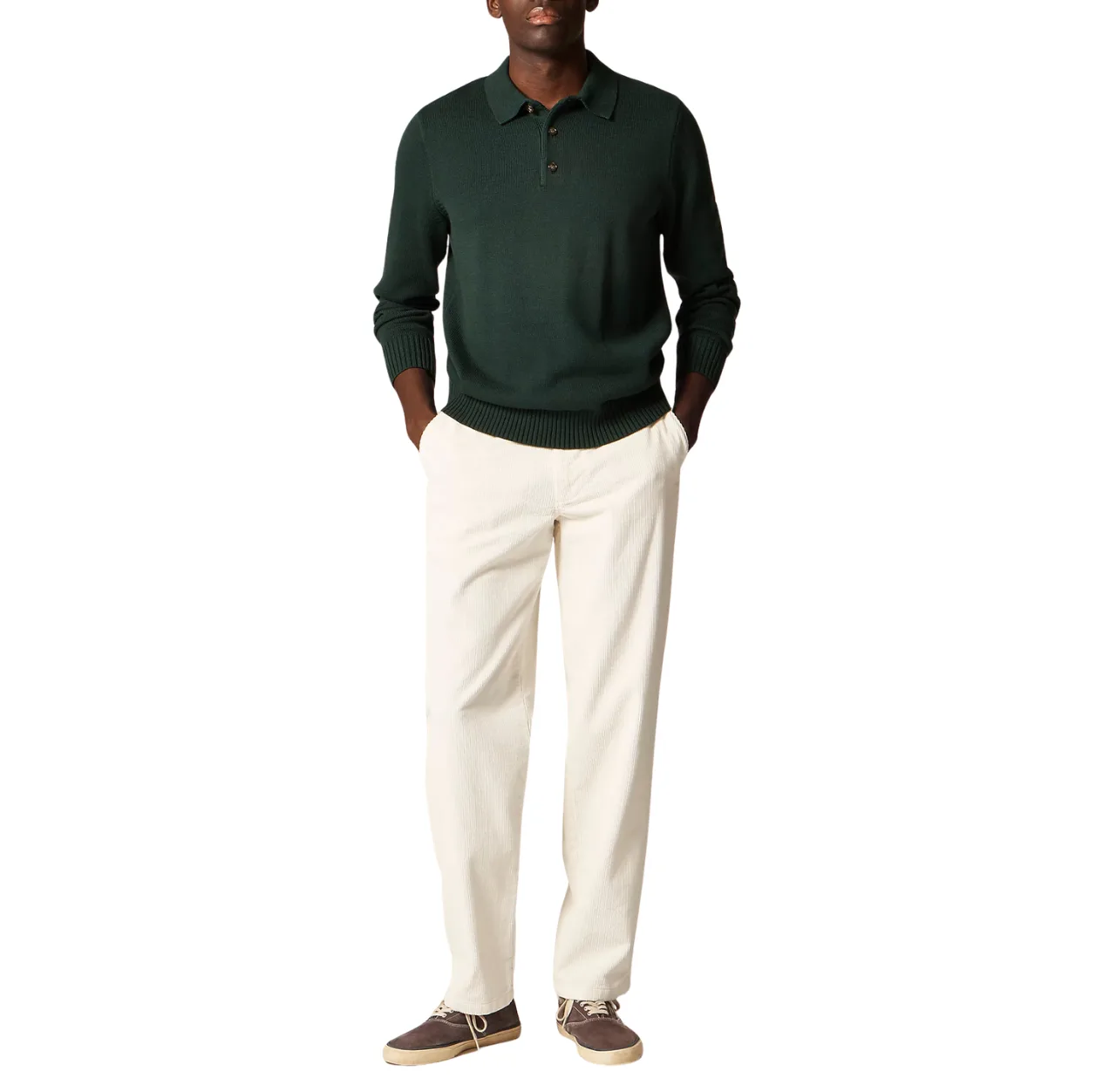 Man wearing a dark green polo shirt with cream corduroy pants and brown canvas sneakers, standing casually with both hands in his pants pockets.