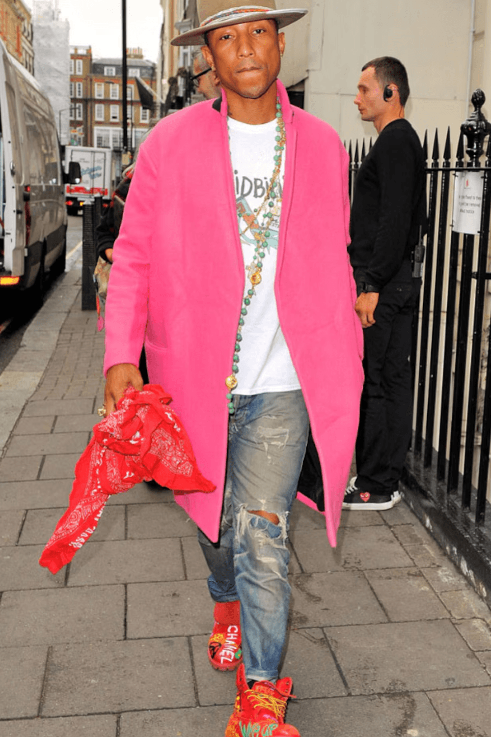 Pharrell Williams wearing a brown hat, white T-shirt, pink overcoat, distressed blue jeans, and red shoes, holding a red bandana in his hand.