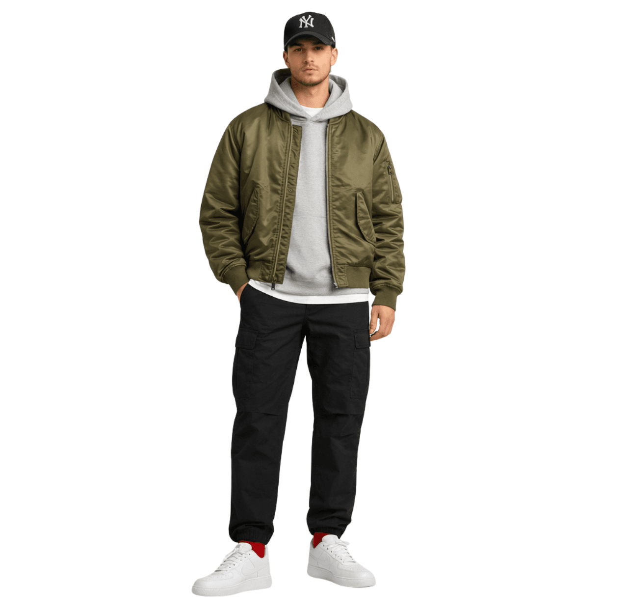Man wearing a black baseball cap, white T-shirt, gray hoodie layered under an olive bomber jacket, relaxed black cargo pants, white sneakers and red socks, standing with one hand in his pocket.