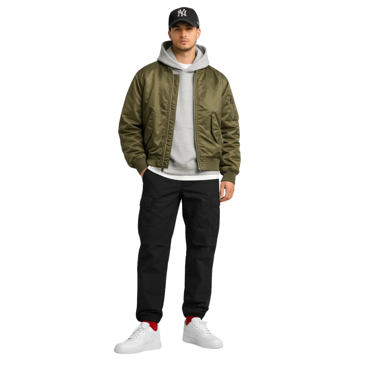Man wearing a black baseball cap, white T-shirt, gray hoodie layered under an olive bomber jacket, relaxed black cargo pants, white sneakers and red socks, standing with one hand in his pocket.