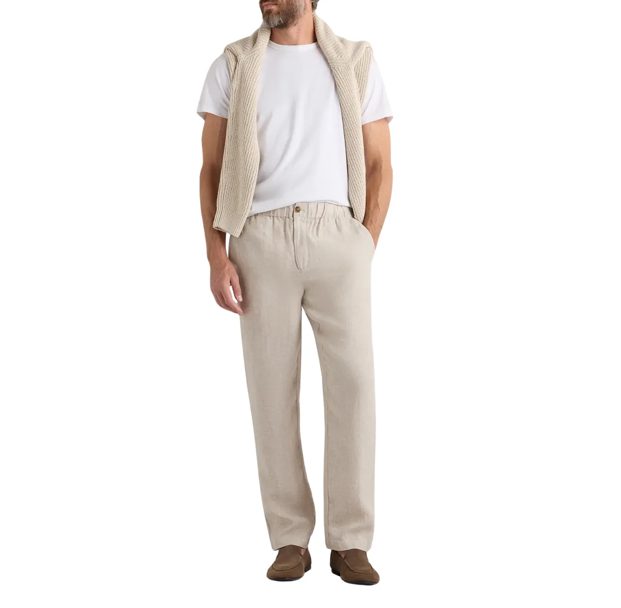 Man wearing beige linen trousers with a white T-shirt and brown slip-on sneakers, optionally layered with a beige sweater, standing with one hand in his pants pocket.