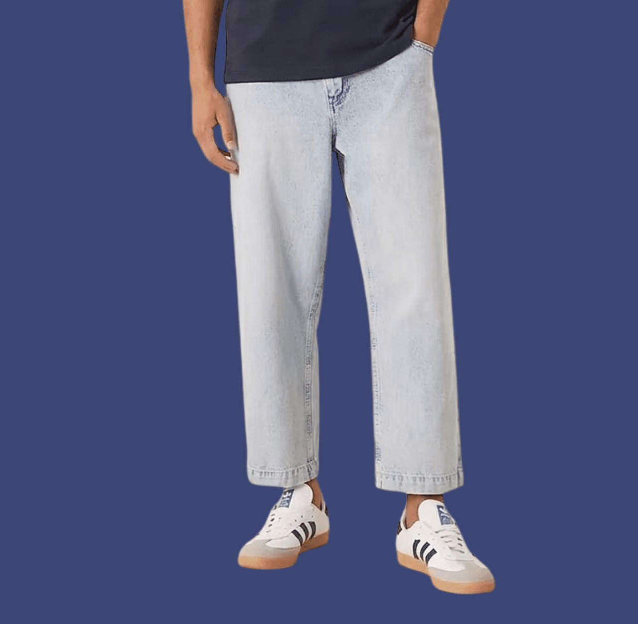 Man wearing blue cropped jeans and white Adidas Samba sneakers, standing casually with one hand in his pocket.