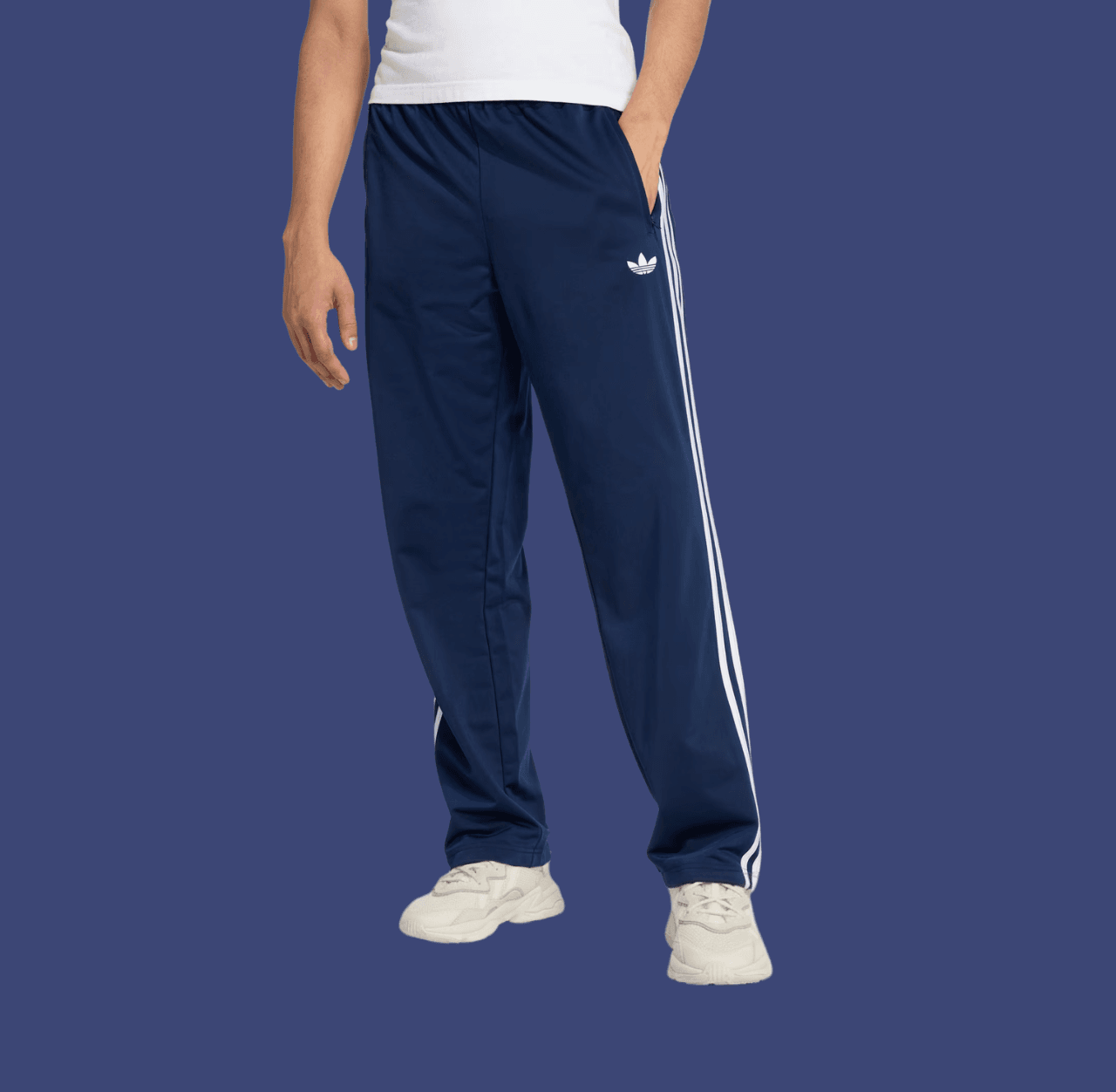 Man in navy relaxed track pants and white chunky sneakers with a hand in pockets, standing casually.
