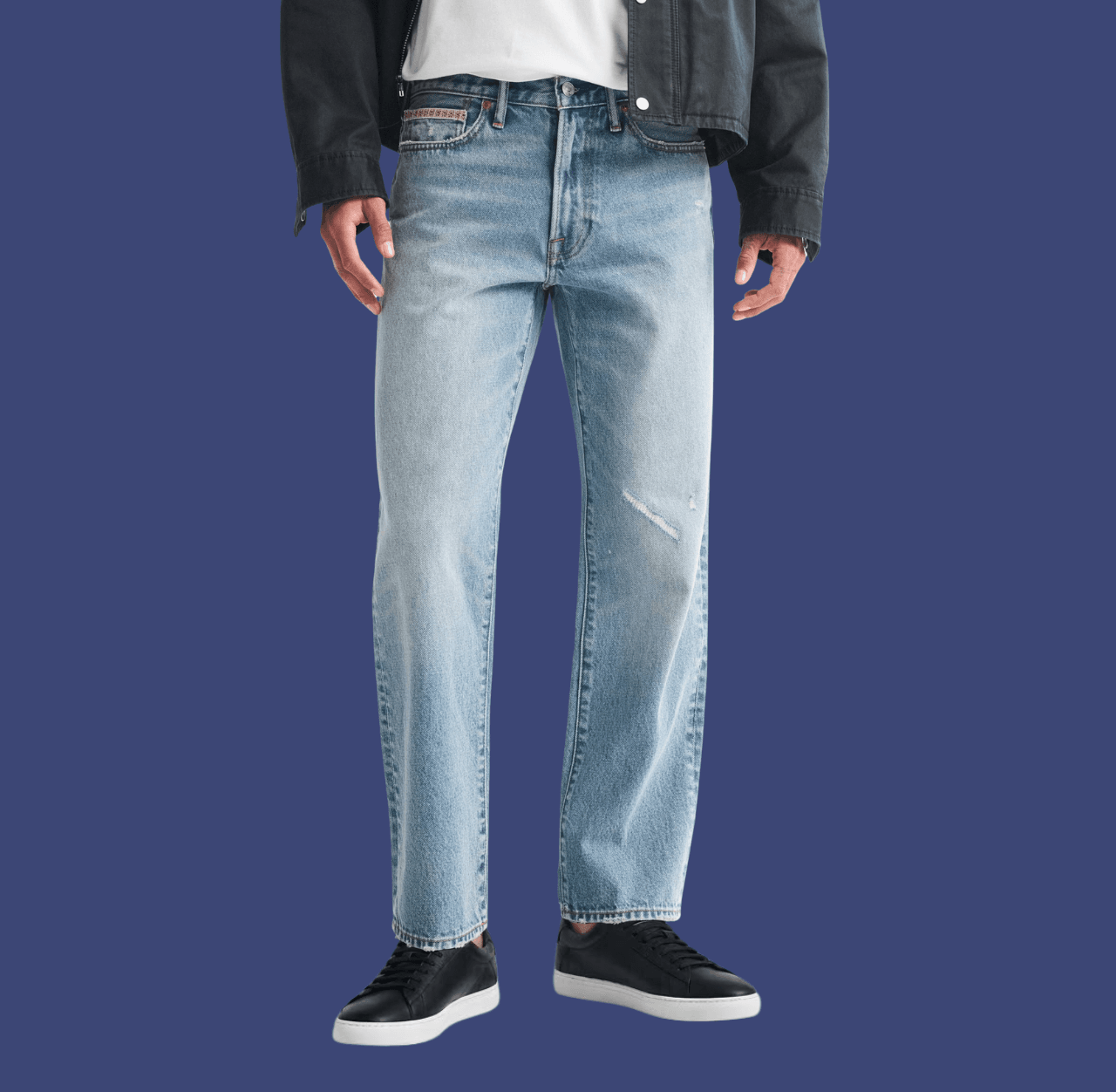 Man wearing blue straight-fit jeans and black low-top sneakers, standing casually with arms relaxed at his sides.