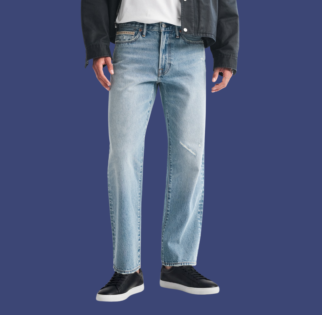 Man wearing blue straight-fit jeans and black low-top sneakers, standing casually with arms relaxed at his sides.