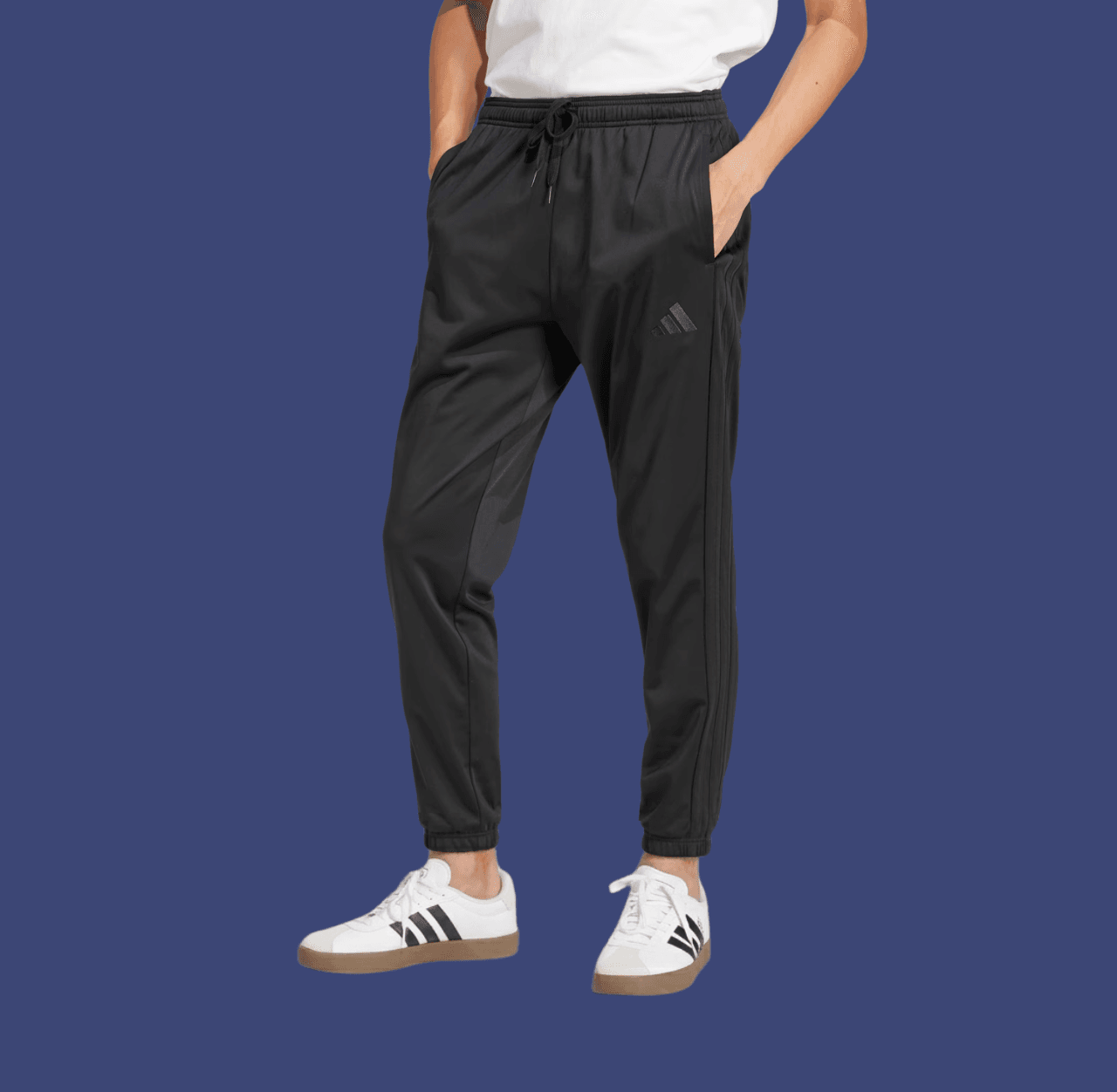 Man in black tapered track pants and white Adidas Samba sneakers with hands in pockets, standing casually.