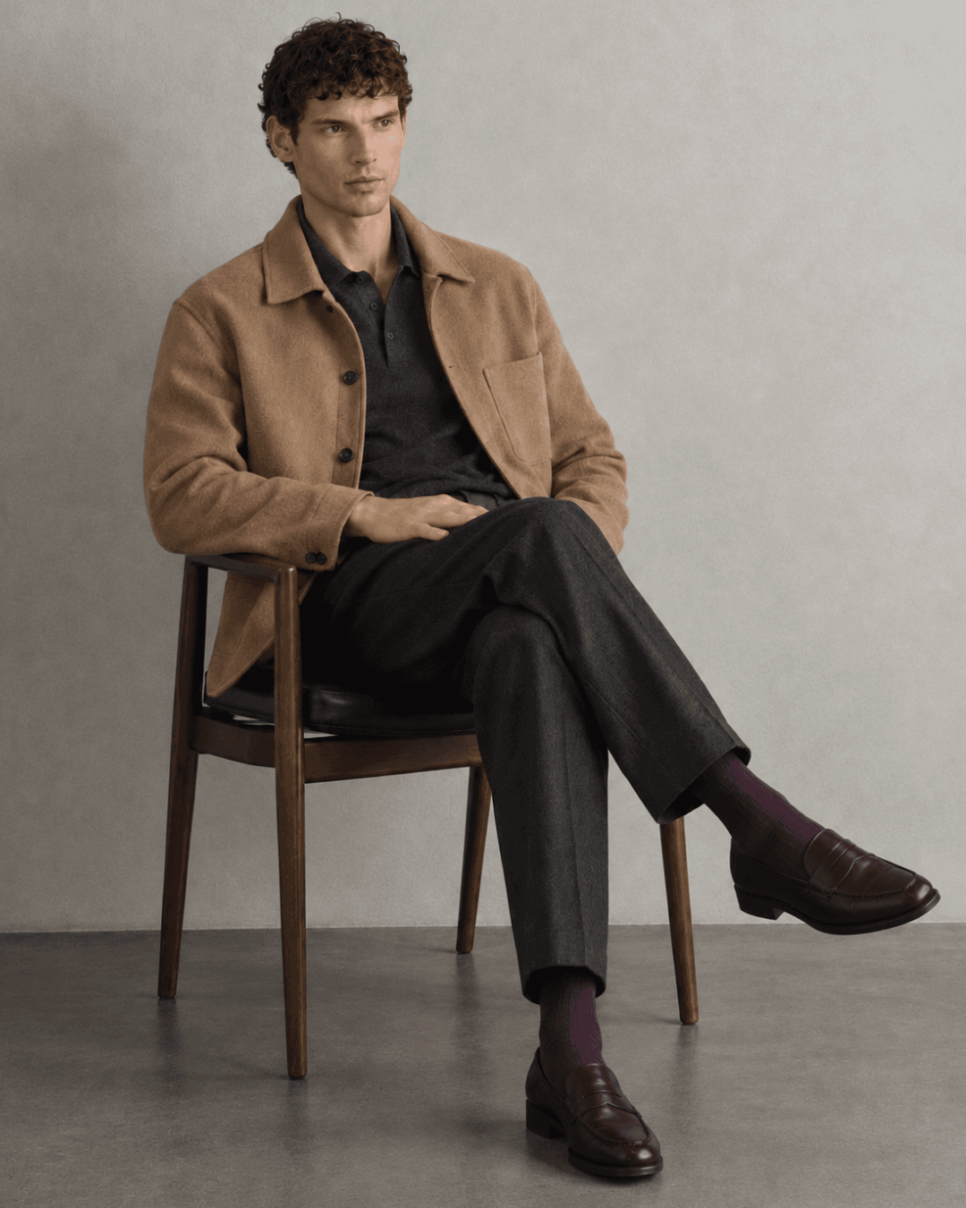 A Man wearing a Camel wool overshirt, a Charcoal knit polo, Dark charcoal pleated trousers, Deep plum socks, and Black leather loafers