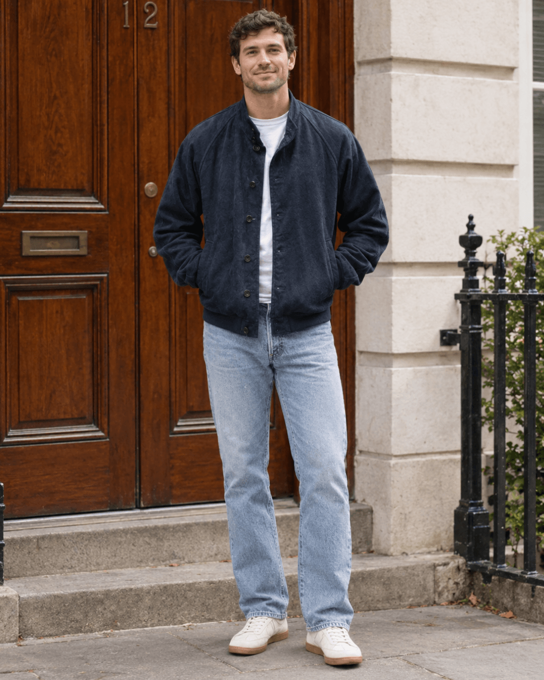 A man wearing a suede bomber jacket, a white tee, blue jeans, and sneakers