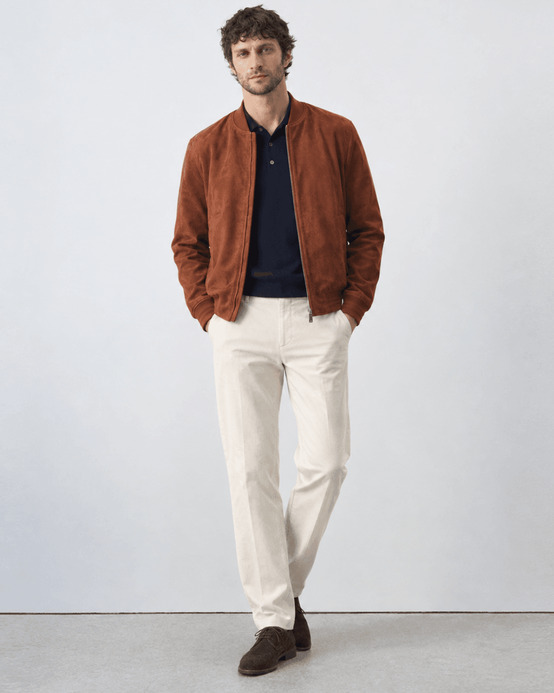 Man wearing a Rust-colored suede bomber, a Navy fine-gauge knit polo, Cream trousers, and Dark brown suede derbies