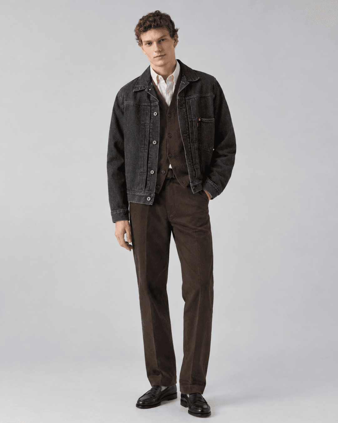 Man wearing a Washed black denim jacket, a Tobacco brown cardigan, a Cream Oxford shirt, Dark brown trousers, and Black loafers