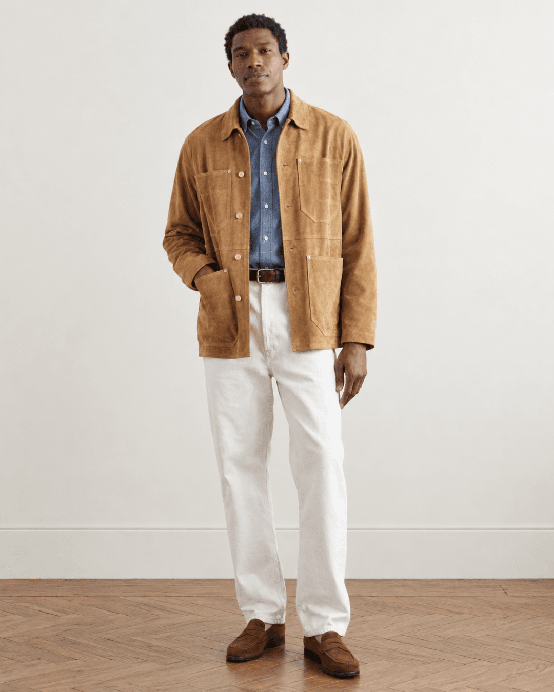 Man wearing a suede chore jacket, a chambray shirt, white jeans, ans suede loafers